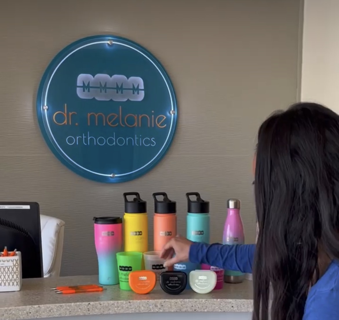 Front desk display of Dr. Melanie Orthodontics branded water bottles, cups, pens, and retainer cases.